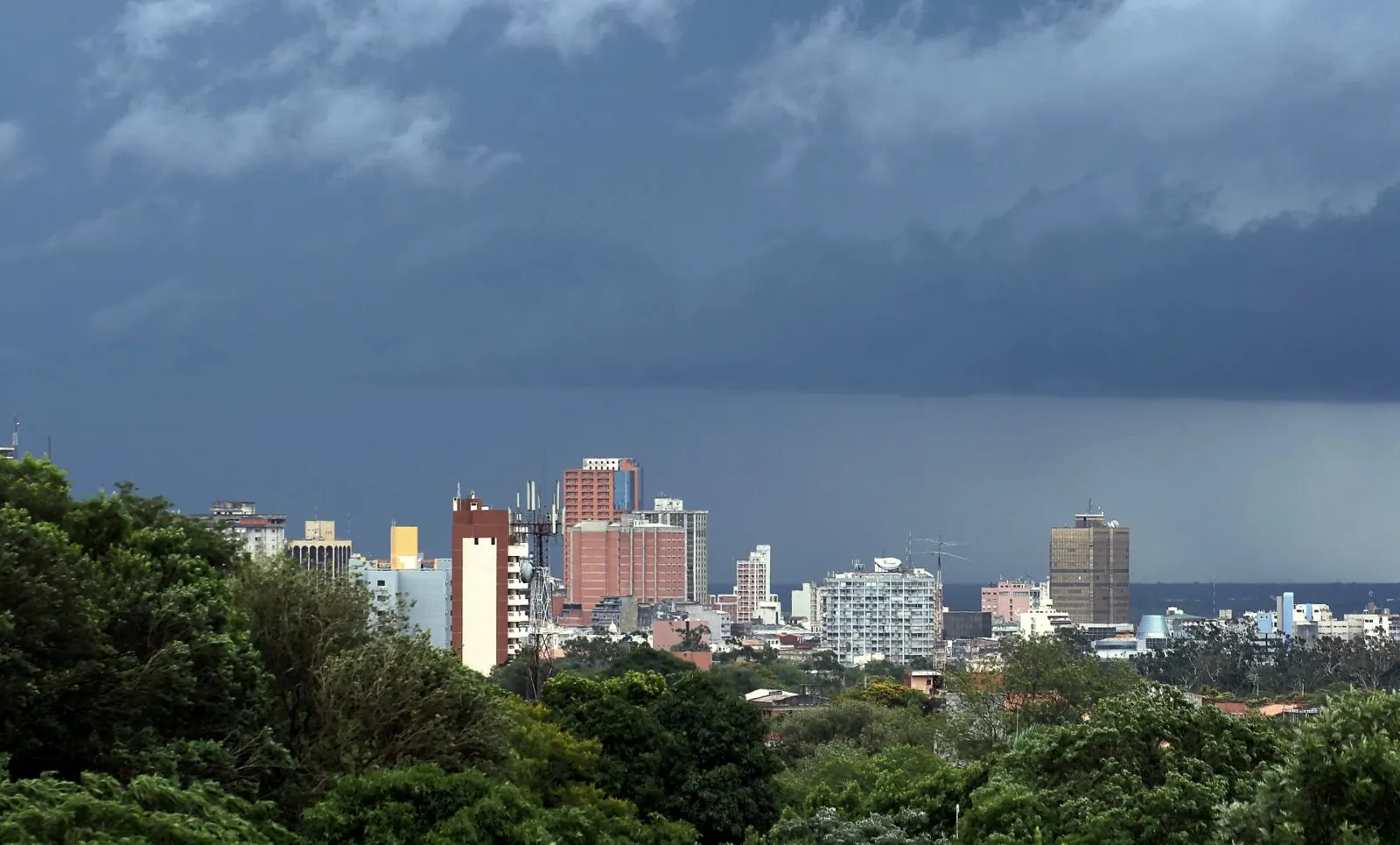 Dark storm clouds and falling rain over the Asunción skyline, with trees in the foreground.