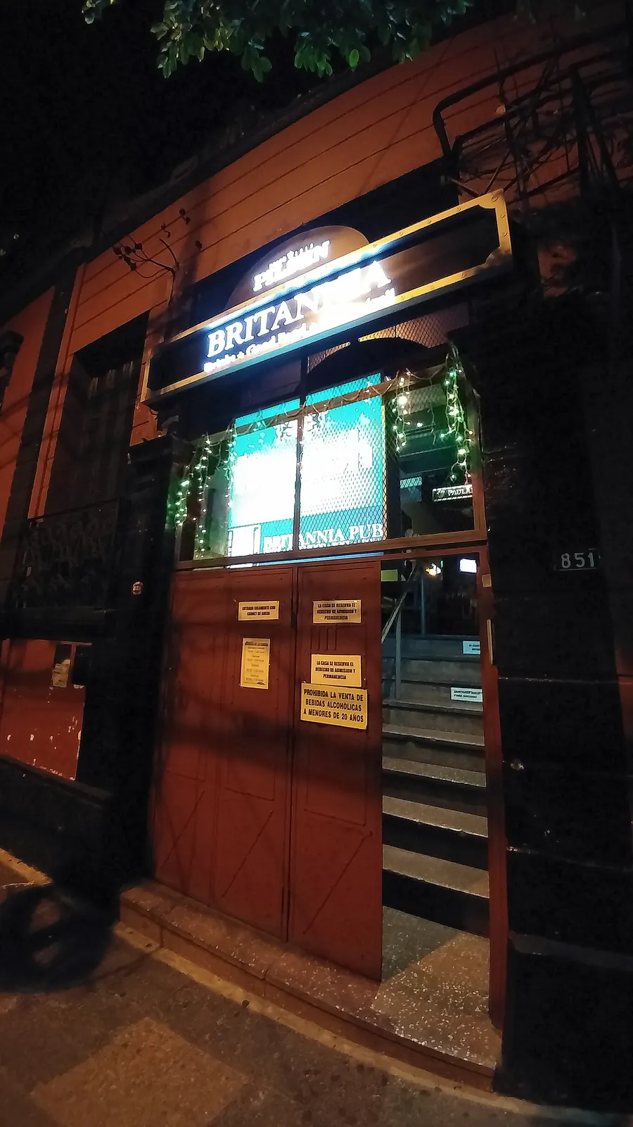 El frente del Britannia Pub de noche, con el cartel iluminado sobre una puerta roja abierta y una escalera que sube.