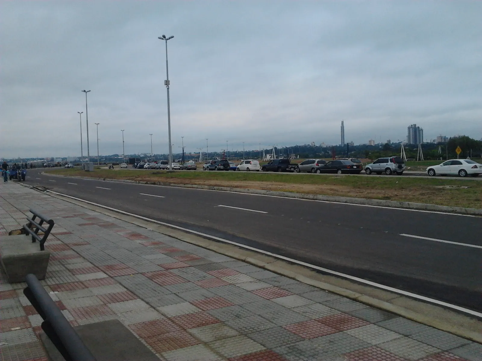 Un paseo pavimentado junto al río, con un banco al lado de la avenida Costanera, faroles y autos estacionados bajo un cielo gris