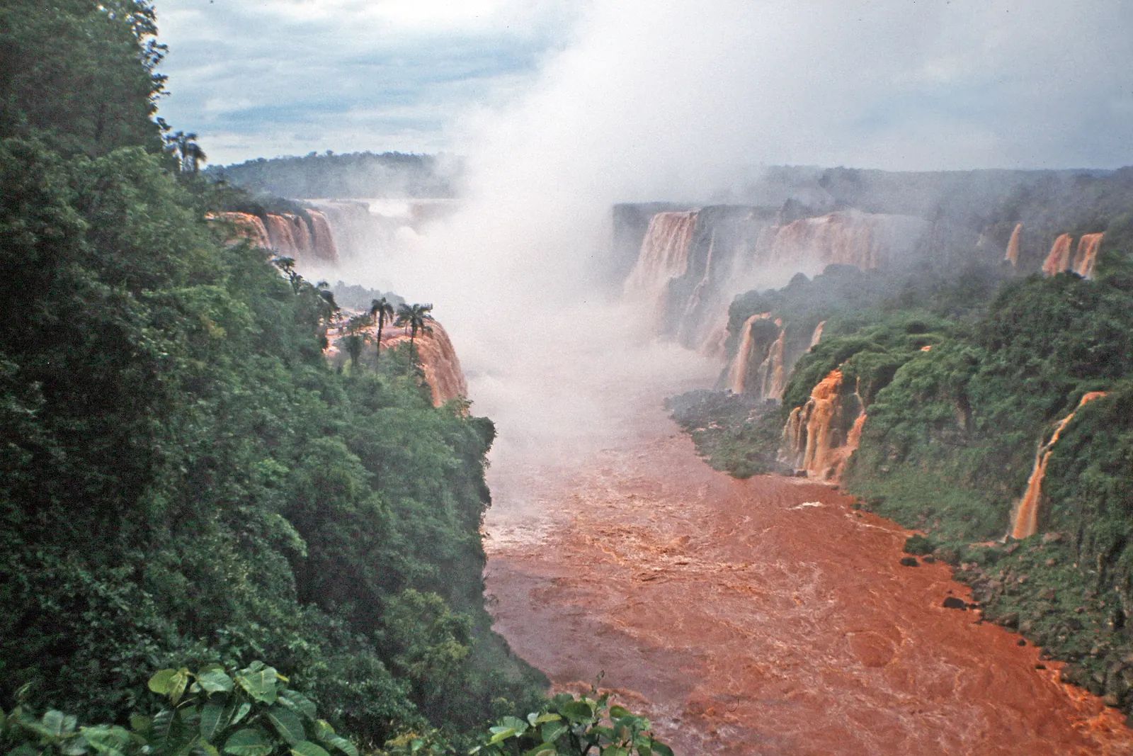 Several waterfalls pouring into a misty gorge above a brown, flooded river, with forest on both banks