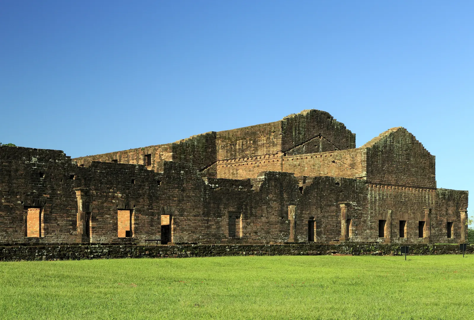 Roofless stone walls with rows of windows and doorways on the south side of the unfinished church at Jesús de Tavarangue, a lawn in front