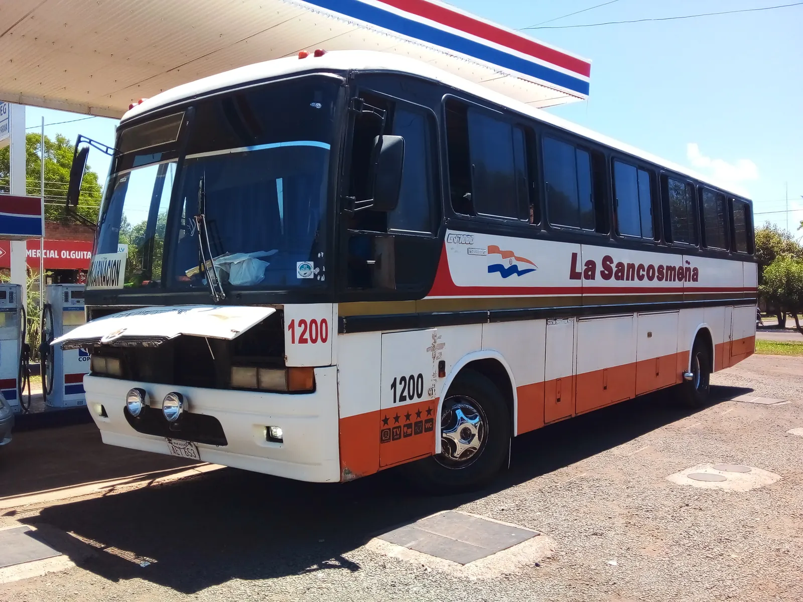A white and orange local bus lettered La Sancosmeña parked at a fuel station