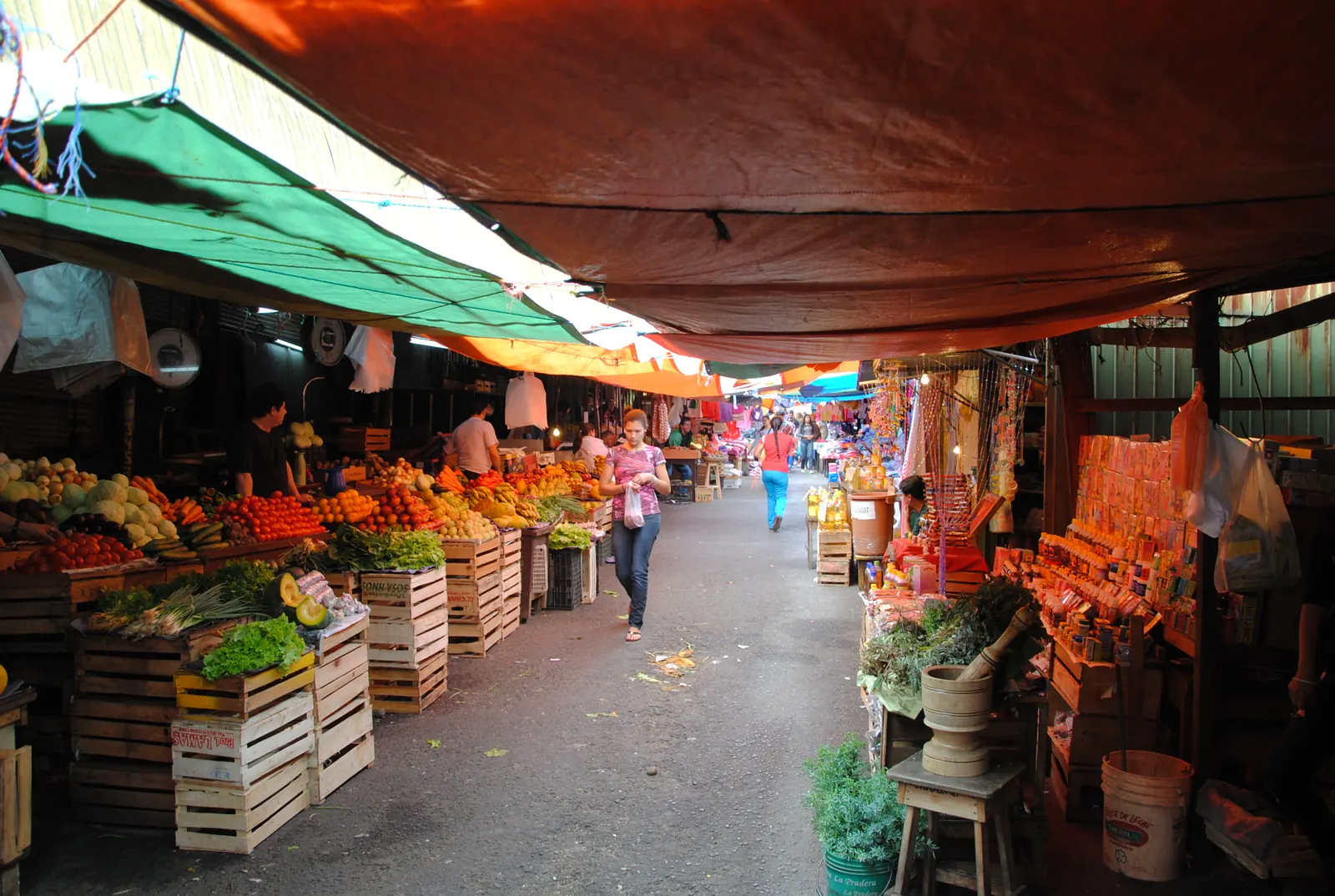 Aisle at Mercado 4 lined with crates of fruit and vegetables under green and orange awnings, with a wooden mortar and bundles of herbs on the right