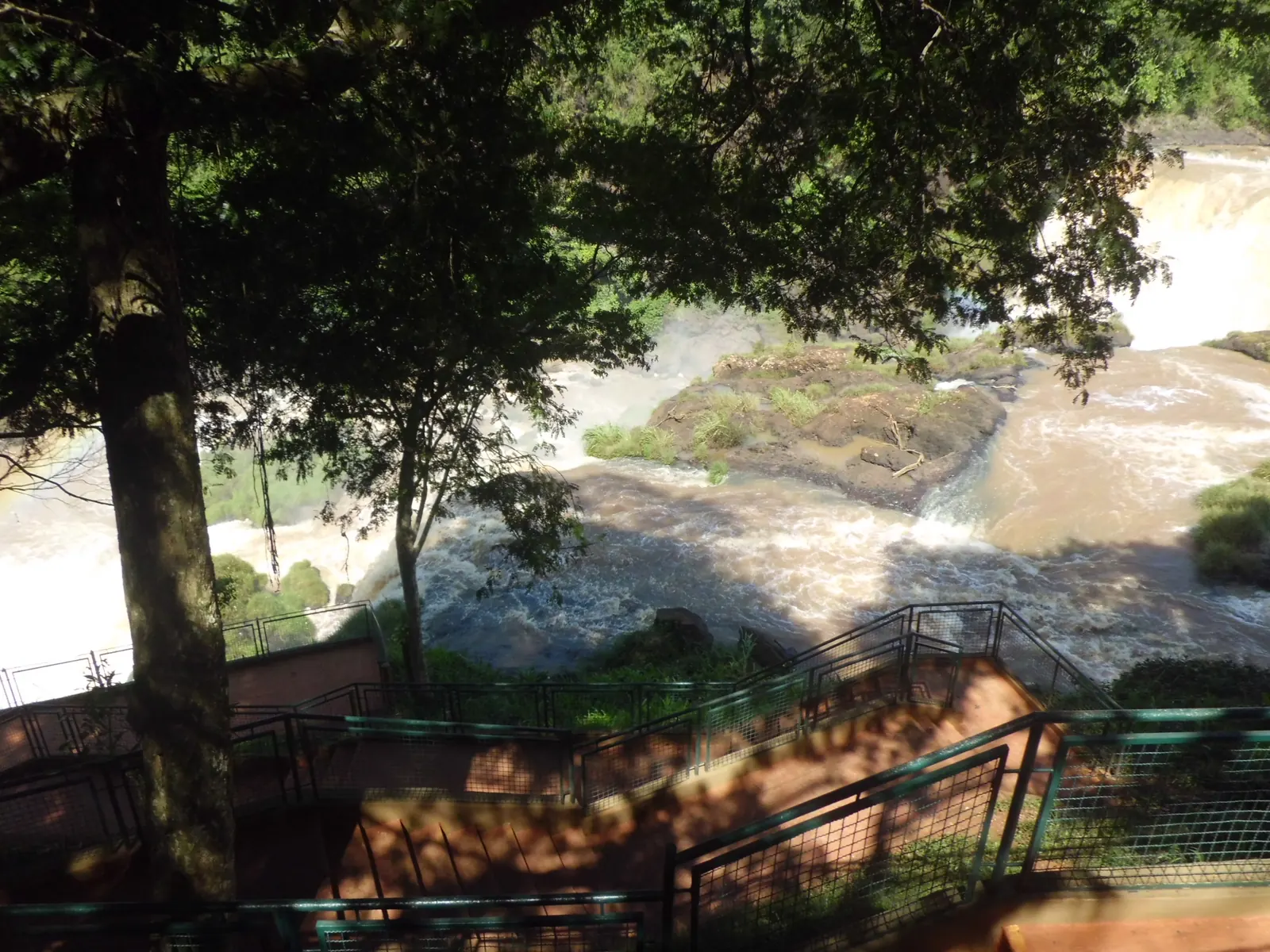 Fenced stairways under trees leading down beside rapids, with a waterfall at the top right