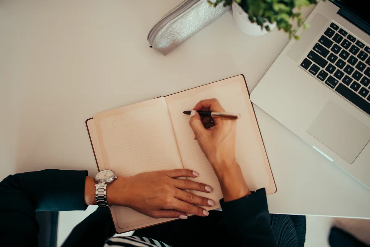 Manos escribiendo en un cuaderno abierto junto a una computadora portátil.