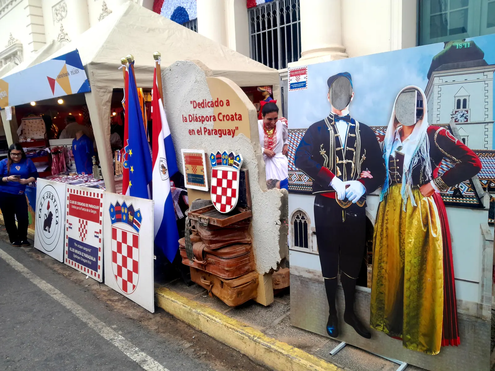 A fair stall with Croatian and Paraguayan flags, old suitcases and a photo board of two figures in folk dress, set up on a street in front of historic buildings