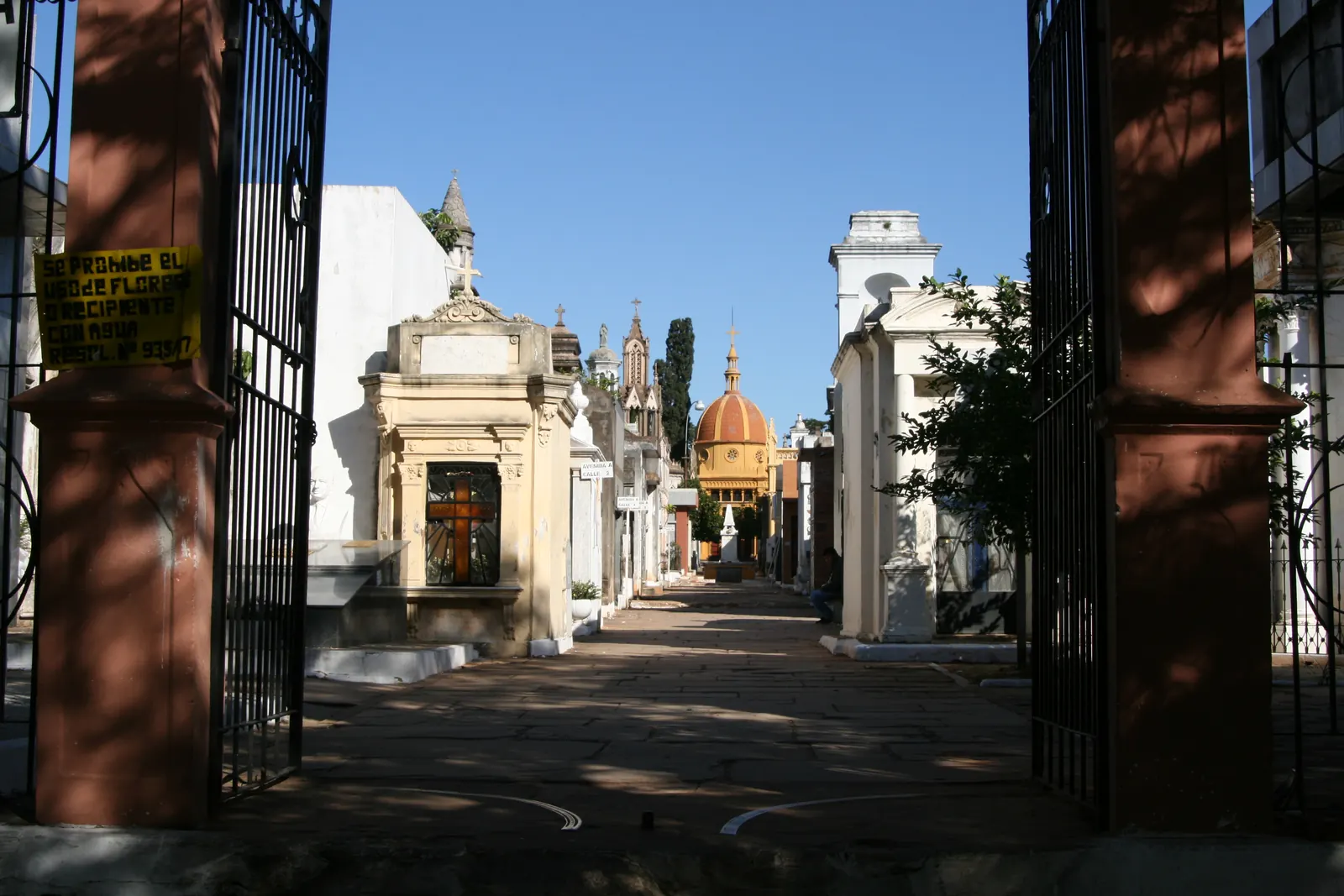 Portones de hierro abiertos que dan a un camino entre tumbas blancas hasta una capilla de cúpula naranja
