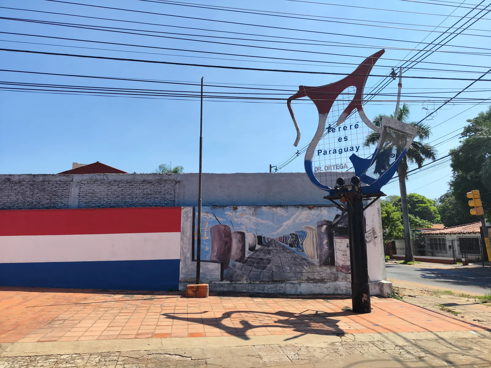 Street sculpture of a tereré jug and guampa reading Tereré es Paraguay, beside a mural of guampas and a Paraguayan flag on a wall.