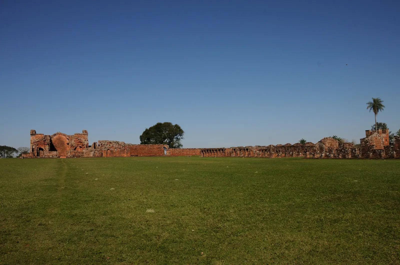 A long line of red sandstone mission ruins across a wide lawn under a blue sky at Trinidad