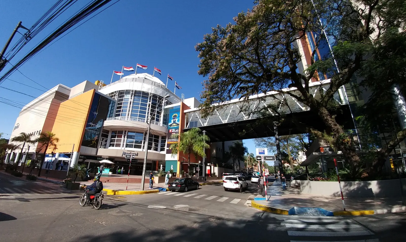 A street crossing in front of a shopping center with a pedestrian bridge in Villa Morra