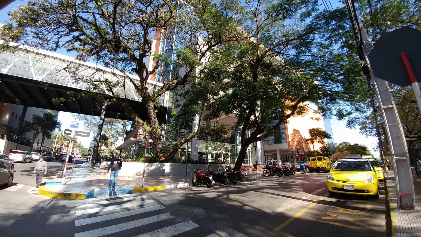 Tree-shaded street corner in Villa Morra with a pedestrian crossing, parked motorbikes and a yellow taxi.
