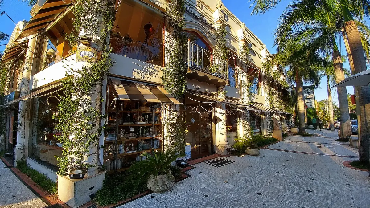 Two-storey shopping arcade with plants on its columns and a wide sidewalk lined with palms.